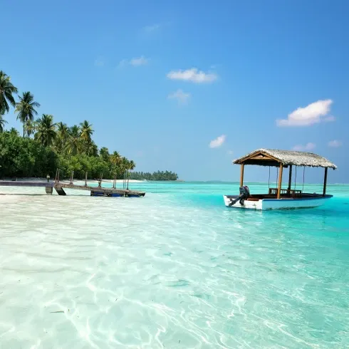 Idyllische Südsee-Kulisse mit einem Boot auf glasklarem Meer, Palmen im Hintergrund und einem Holzsteg am weißen Sandstrand.
