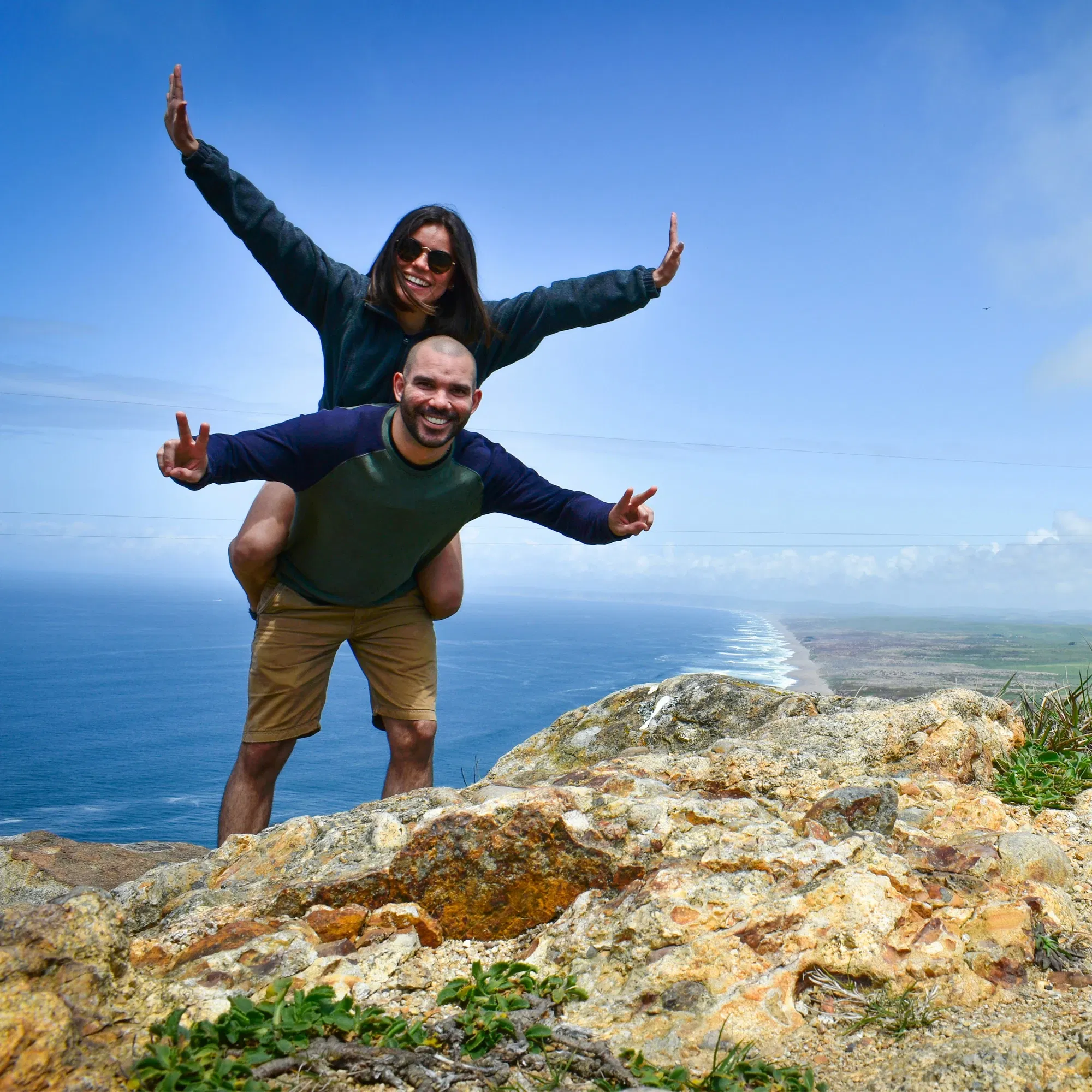 Ein Paar posiert lachend auf einem Felsen mit Blick auf die Küstenlinie, während die Frau auf den Schultern des Mannes sitzt und beide die Arme ausstrecken.