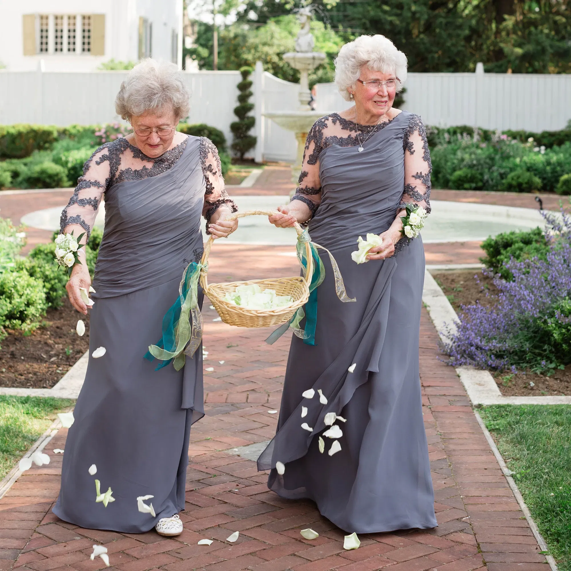 Zwei Großmütter, elegant in grauen Kleidern mit Spitzenärmeln gekleidet, streuen Blütenblätter aus einem Korb. Sie laufen einen Ziegelweg entlang in einem liebevoll gestalteten Garten und zaubern den Hochzeitsgästen ein Lächeln ins Gesicht.