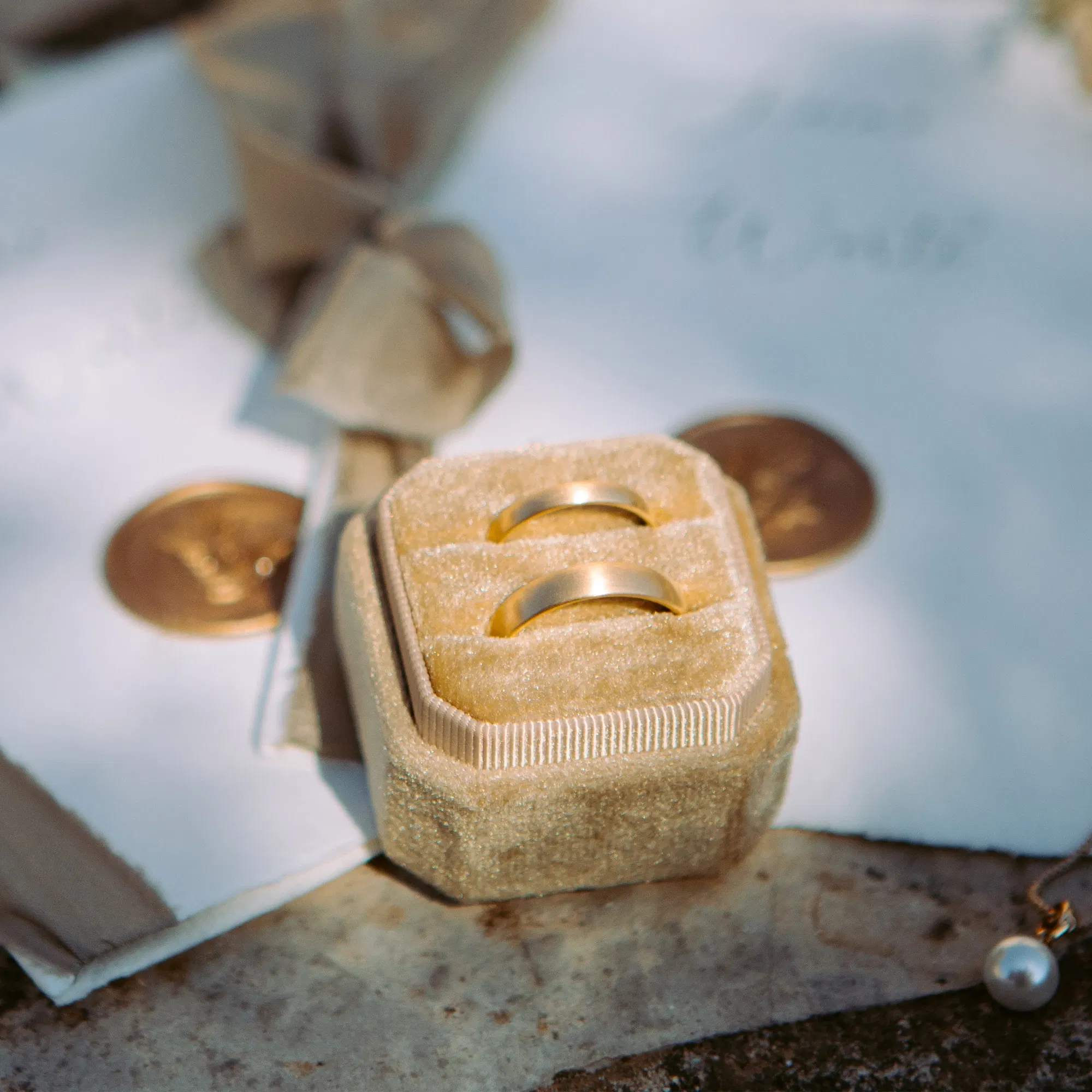 Zwei schlichte goldene Trauringe in einer samtigen Box, arrangiert auf einer Hochzeitsdekoration mit Bändern und Münzen im Hintergrund.