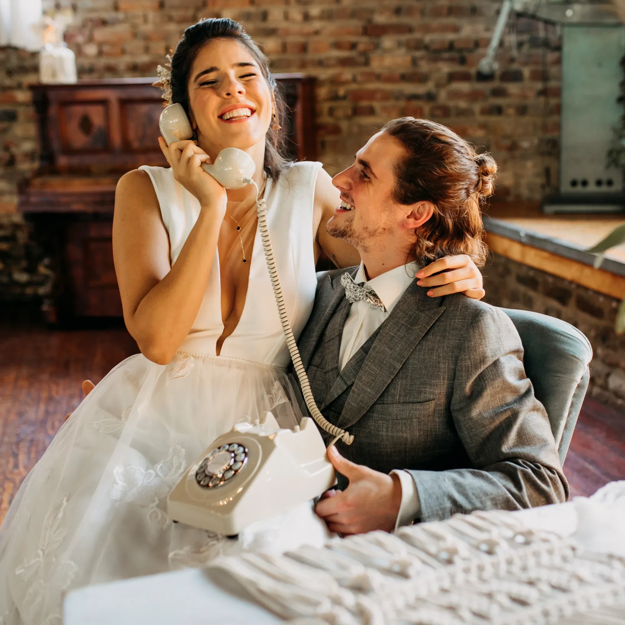 Brautpaar sitzt lachend an einem Retro-Telefon bei der Hochzeit, die Braut hält den Hörer und strahlt vor Freude, der Bräutigam schaut sie liebevoll an.