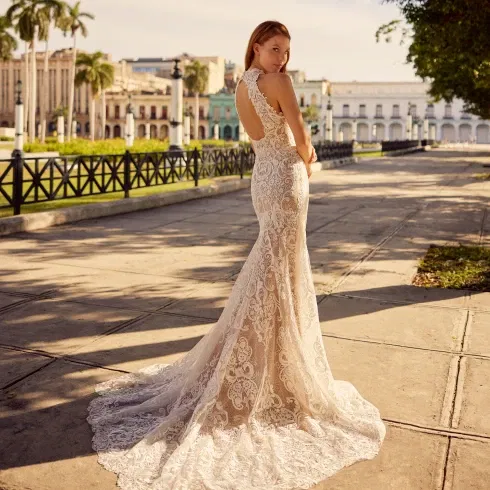 Eine Braut in einem figurbetonten, eleganten Spitzenkleid mit Schleppe und offenem Rückenausschnitt, posierend auf einer sonnigen Promenade.