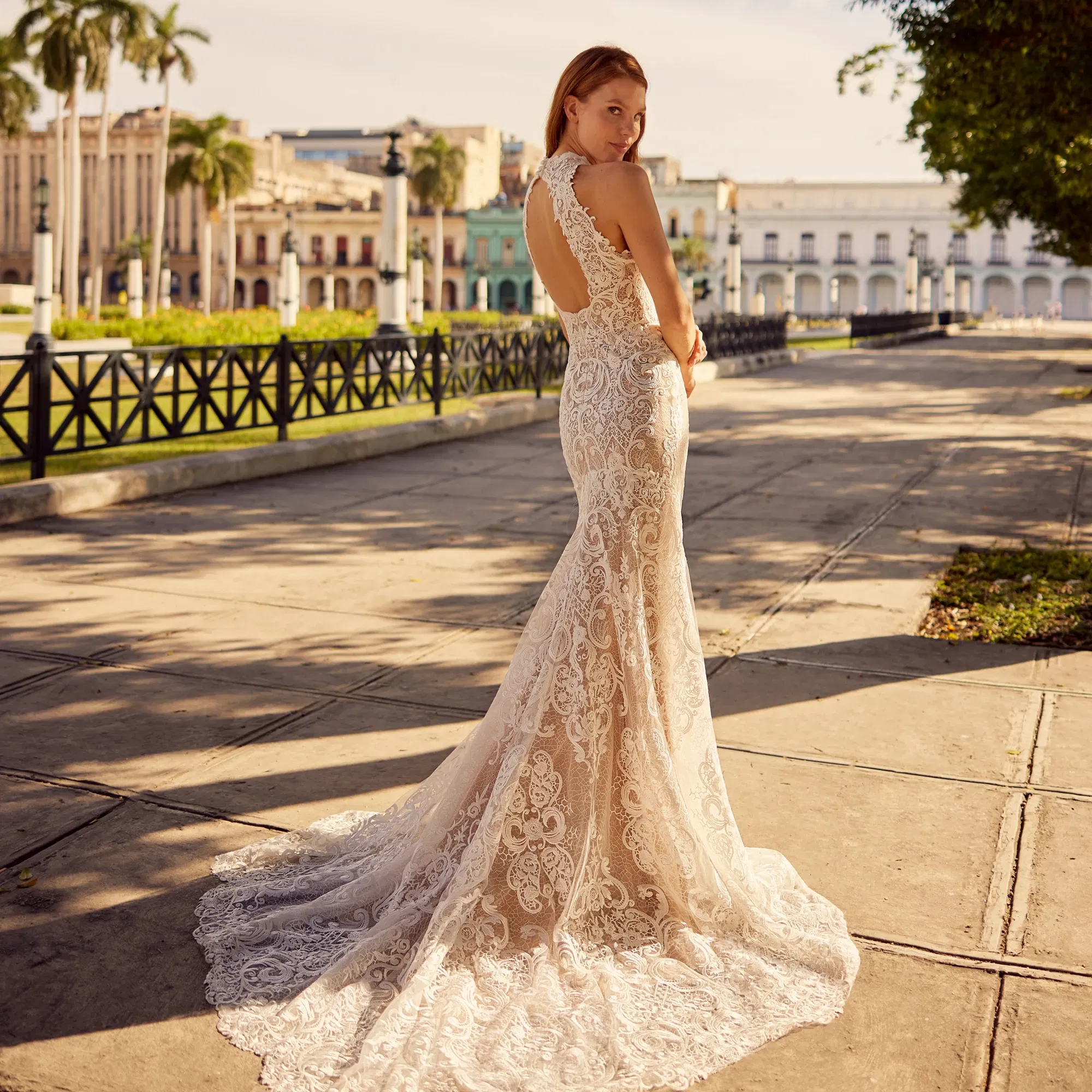 Eine Braut in einem figurbetonten, eleganten Spitzenkleid mit Schleppe und offenem Rückenausschnitt, posierend auf einer sonnigen Promenade.