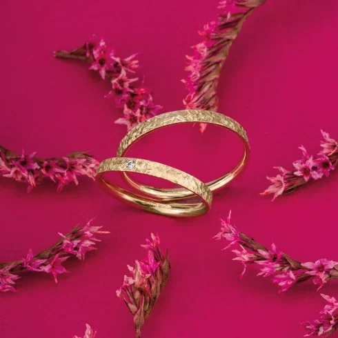 Goldene Eheringe mit filigraner Struktur, dekoriert mit pinken Blüten auf einem kräftig pinken Hintergrund.