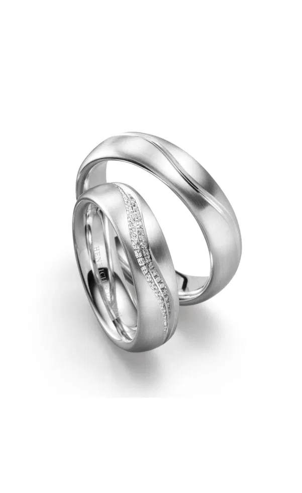 Zwei ineinander stehende Trauringe aus mattiertem Weißgold oder Platin, mit sanft geschwungener Linienführung; ein Ring ist mit einer feinen Diamantreihe entlang der geschwungenen Fuge besetzt.