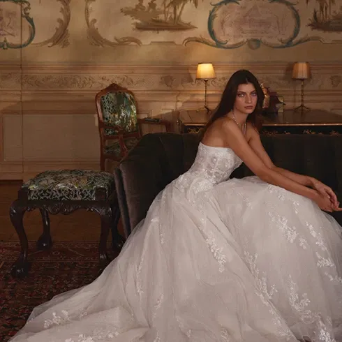 Romantisches Brautkleid mit floralen Stickereien und ausladender Schleppe, sitzend vor einer kunstvoll bemalten Wandkulisse.