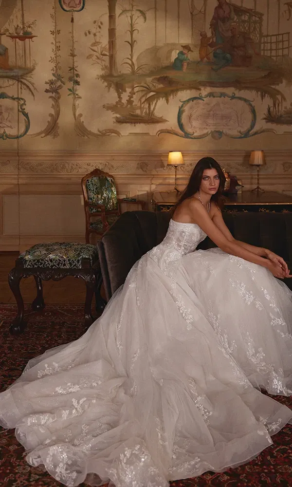 Romantisches Brautkleid mit floralen Stickereien und ausladender Schleppe, sitzend vor einer kunstvoll bemalten Wandkulisse.