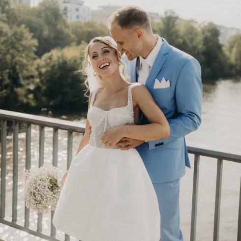 Ein Brautpaar steht lächelnd auf einer Brücke, die Braut in einem modernen weißen Kleid mit Schleier und Blumenstrauß, der Bräutigam in einem hellblauen Anzug.