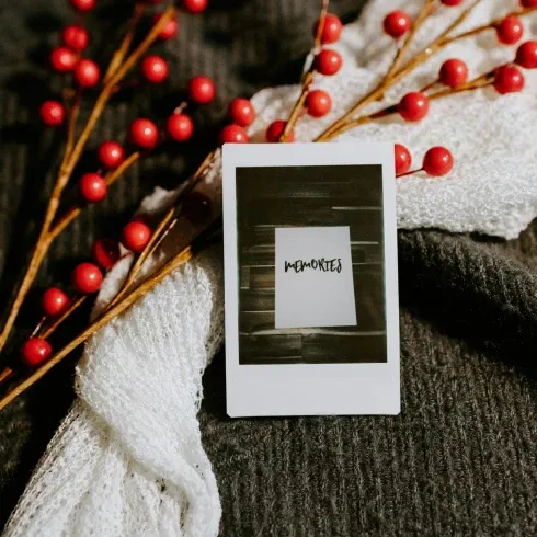 Quadratisches Polaroid mit der Aufschrift "Memories", umrahmt von roten Beerenzweigen und einem weißen Schal auf einer dunklen Decke.