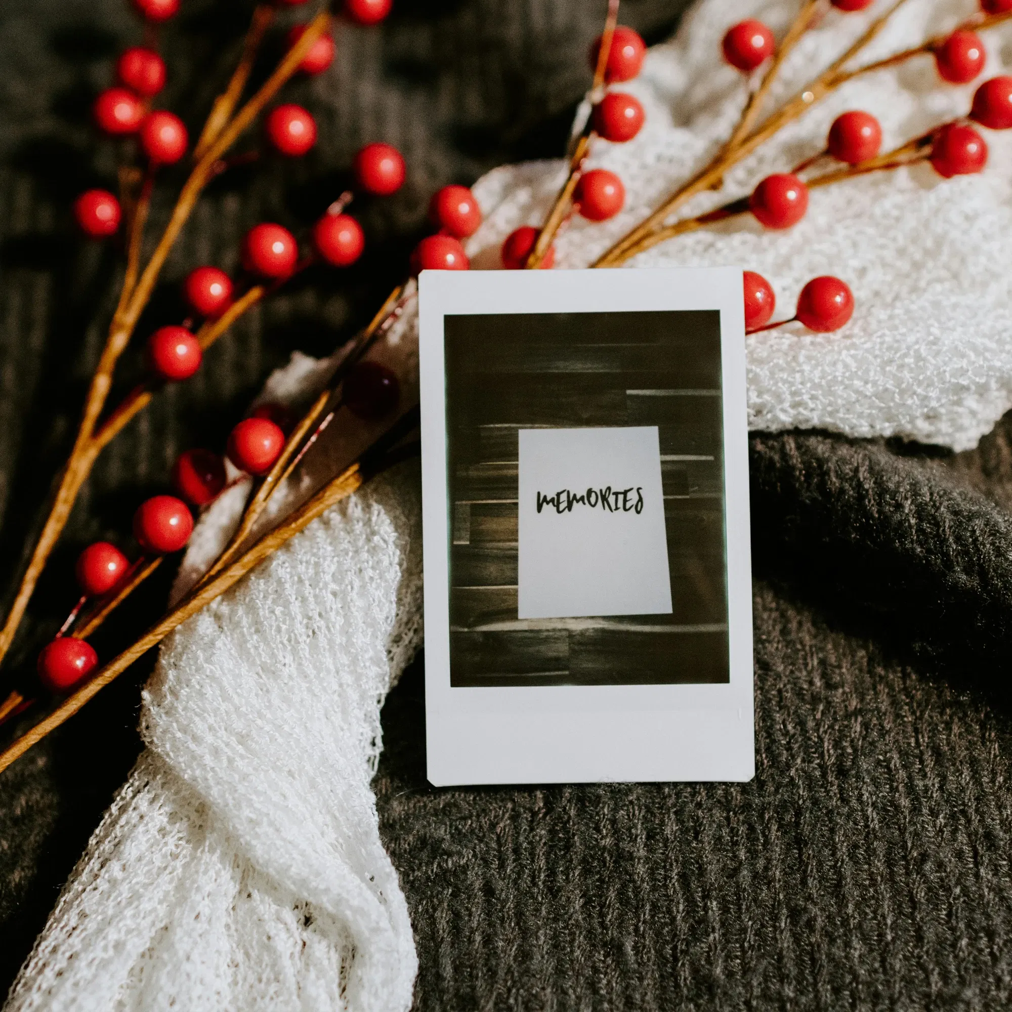Quadratisches Polaroid mit der Aufschrift "Memories", umrahmt von roten Beerenzweigen und einem weißen Schal auf einer dunklen Decke.