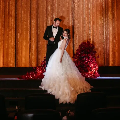 Ein elegantes Brautpaar posiert auf der Bühne eines Kinos vor einem goldenen Vorhang. Die Braut trägt ein voluminöses weißes Kleid, der Bräutigam einen klassischen schwarzen Anzug. Im Hintergrund sind leuchtend rote Rosen dekorativ arrangiert.