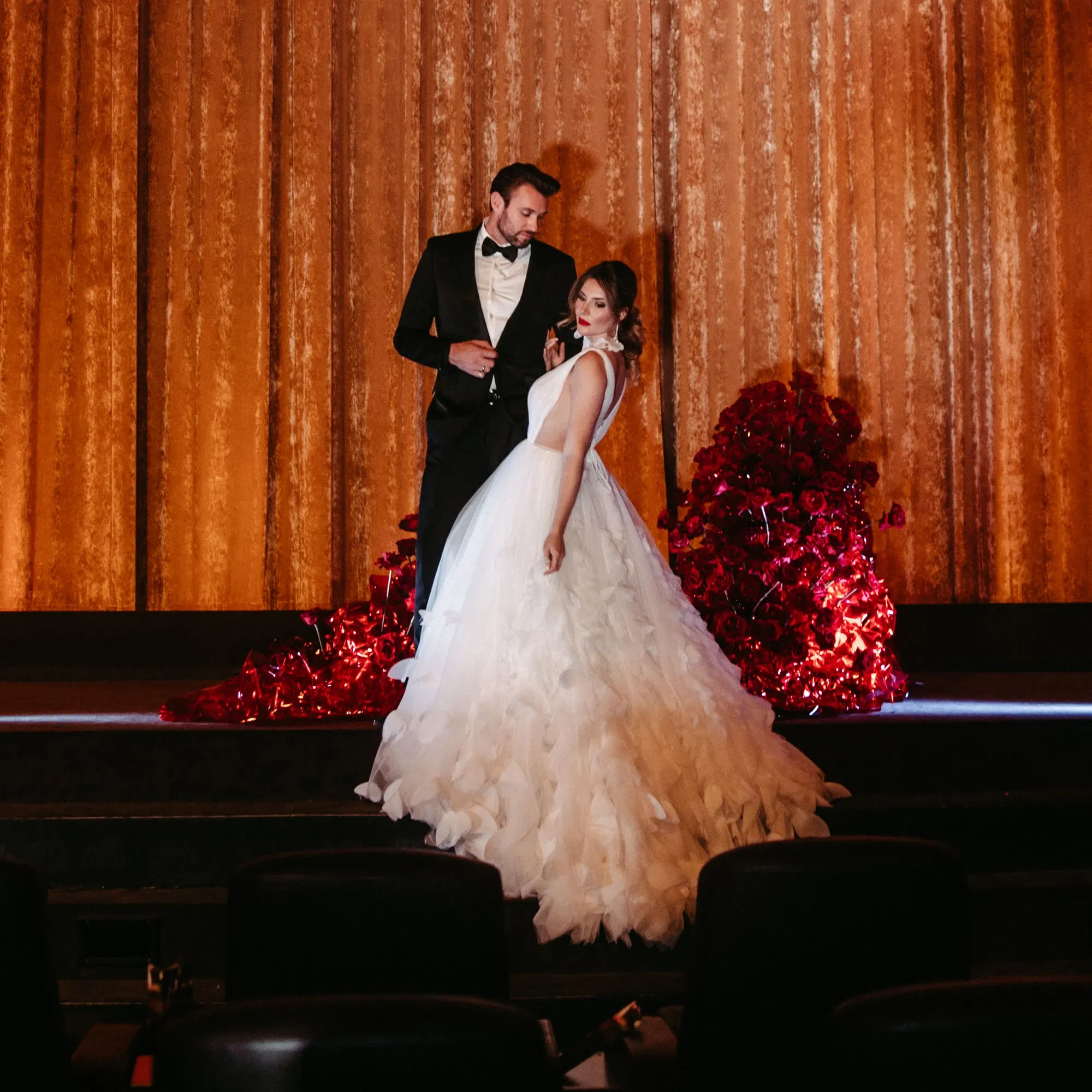Ein elegantes Brautpaar posiert auf der Bühne eines Kinos vor einem goldenen Vorhang. Die Braut trägt ein voluminöses weißes Kleid, der Bräutigam einen klassischen schwarzen Anzug. Im Hintergrund sind leuchtend rote Rosen dekorativ arrangiert.