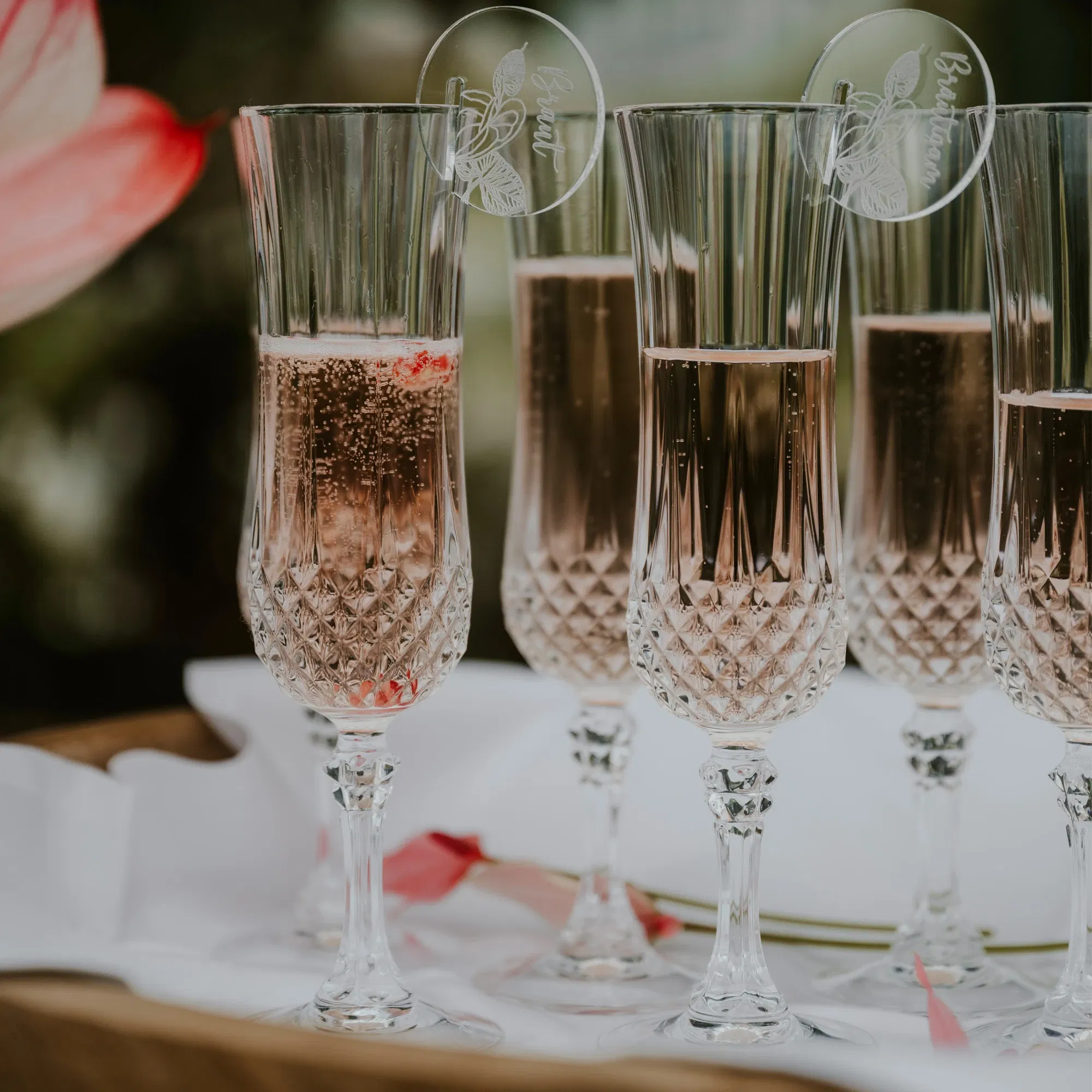 Kristallgläser mit Rosé-Sekt, dekoriert mit individuellen Acryl-Anhängern, stehen stilvoll auf einer Hochzeitstafel und sorgen für einen edlen Empfang.