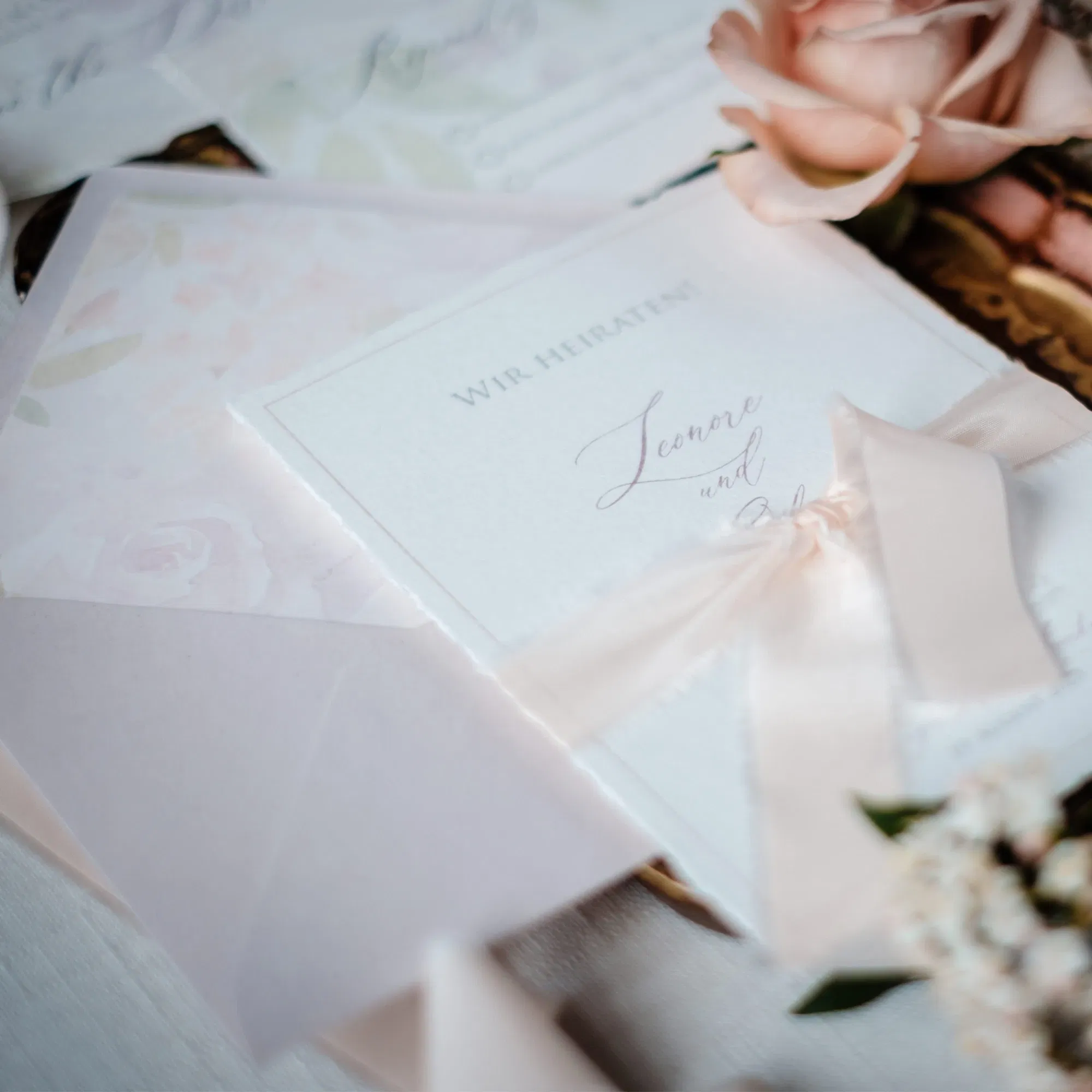 Elegante Hochzeitseinladung mit floralen Details und zartem Seidenband in Rosa, umgeben von romantischen Accessoires.