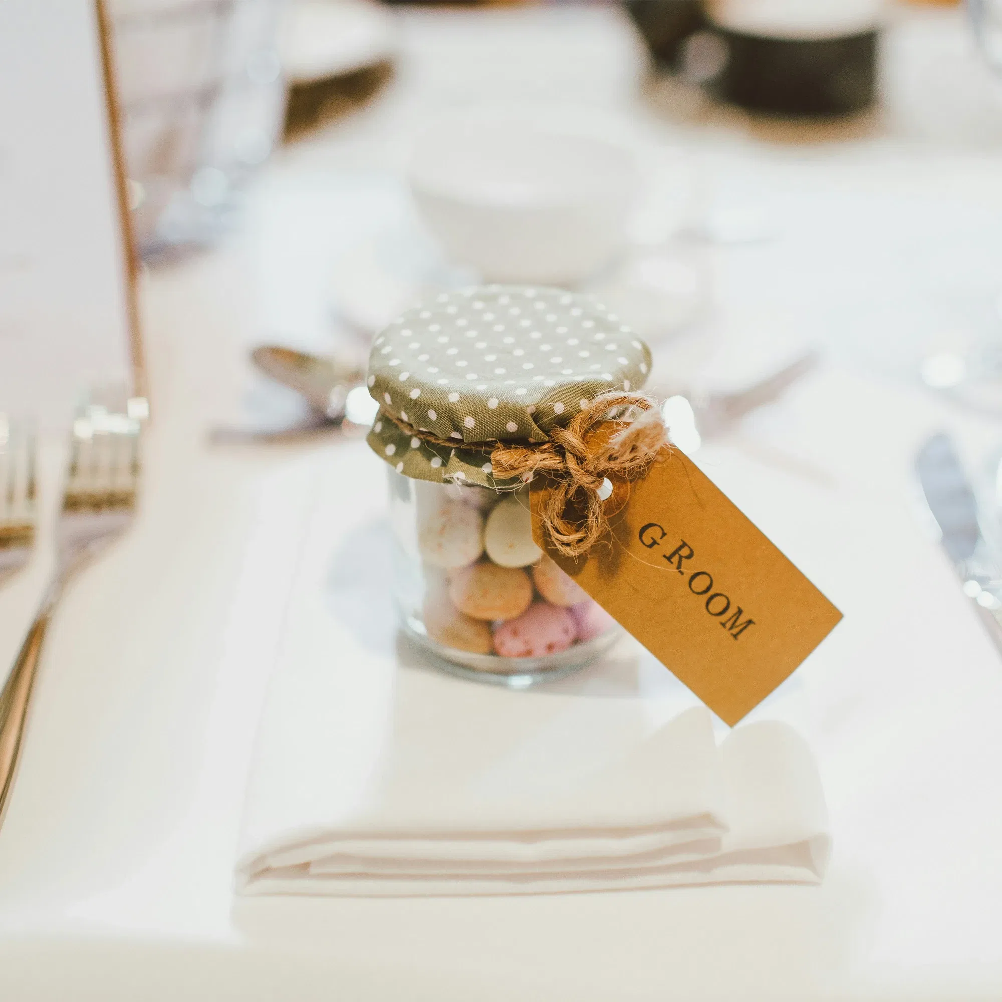 Kleines Glas mit Schokodragees als Gastgeschenk, dekoriert mit gepunktetem Stoffdeckel und Juteschnur, beschriftet mit einem Anhänger ‚Groom‘.