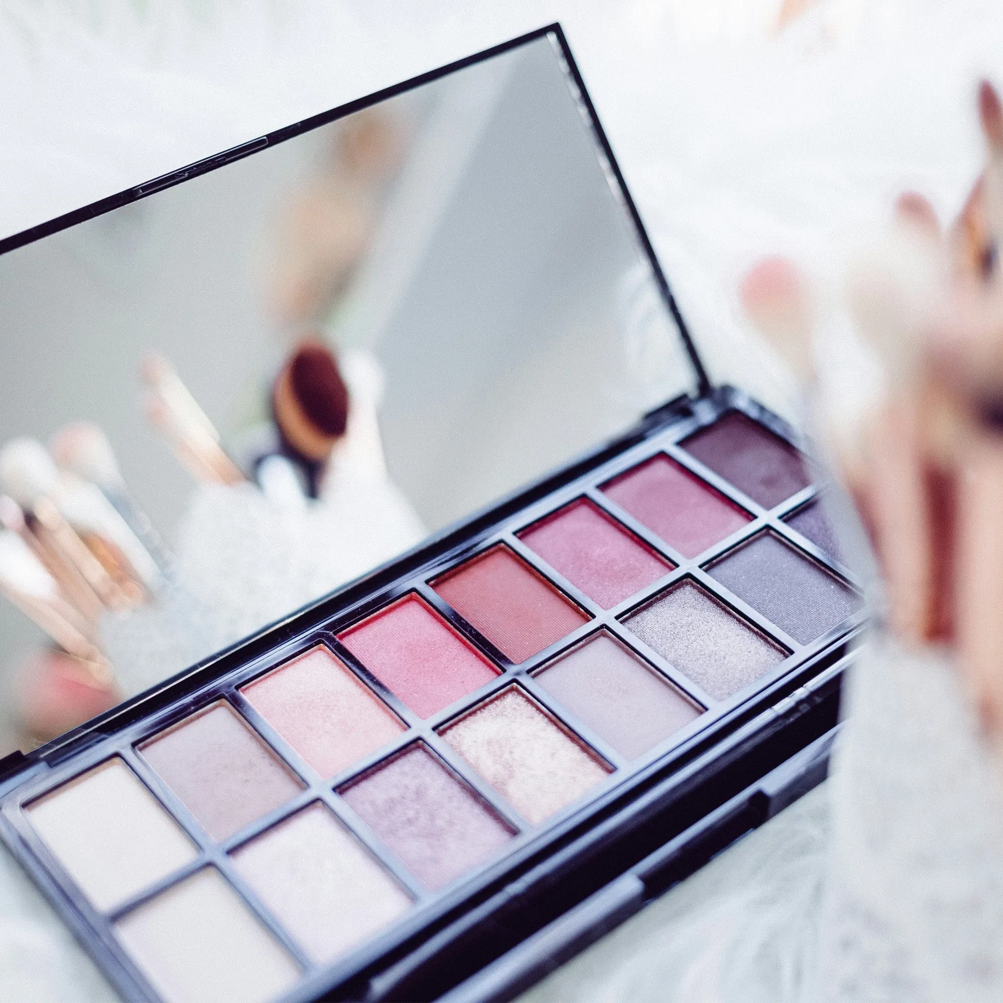 Nahaufnahme einer offenen Lidschattenpalette mit warmen Rosé- und Brauntönen, umgeben von Make-up-Pinseln in weichem Licht.