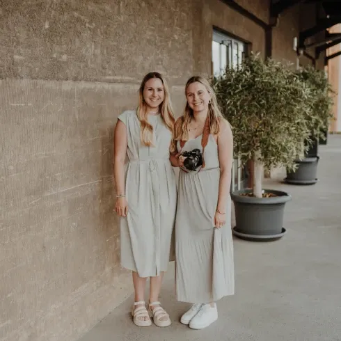 Zwei Frauen stehen nebeneinander vor einer strukturierten Betonwand, lächeln in die Kamera und tragen luftige, helle Kleider. Eine von ihnen hält eine Kamera mit Schultergurt, während im Hintergrund Pflanzen in großen Töpfen eine moderne Location ergänzen.