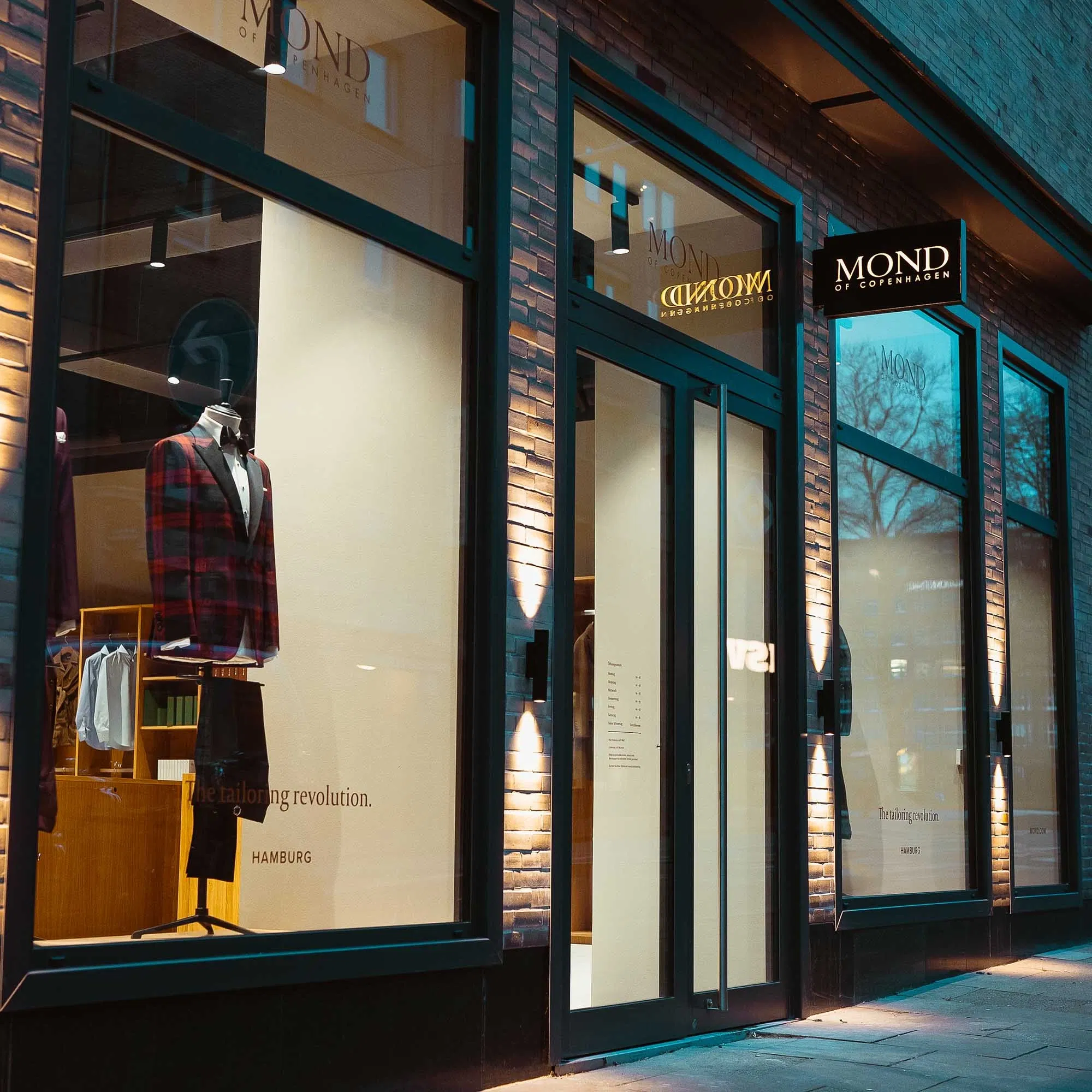 Beleuchtetes Schaufenster eines Herrenausstatters mit elegantem Anzug auf einer Schneiderpuppe und moderner Glasfassade in der Abenddämmerung