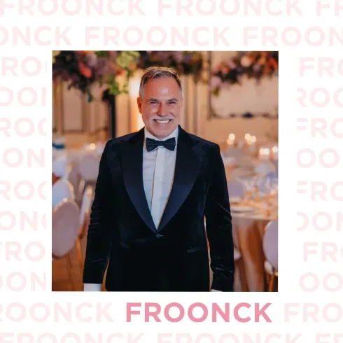Hochzeitsplaner Froonck strahlt in elegantem Smoking vor einer festlich dekorierten Hochzeitslocation mit warmem Kerzenlicht und Blumenarrangements.
