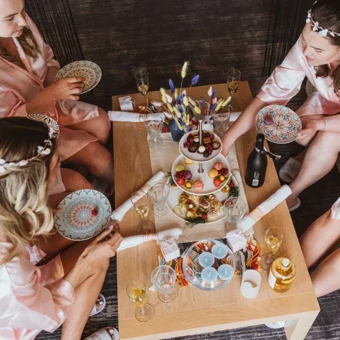 Bridesmaids in rosa Satin-Kimonos genießen ein Champagner-Frühstück mit süßen Leckereien. Eine liebevoll dekorierte Etagere mit Macarons, Obst und Pralinen steht auf dem Tisch, umgeben von stilvollen Gläsern und Blumenarrangements.