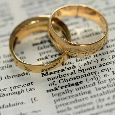Zwei goldene Eheringe liegen auf einer aufgeschlagenen Buchseite mit dem Wort „Marriage“ – Symbol für Ehe, Liebe und rechtliche Bindung.