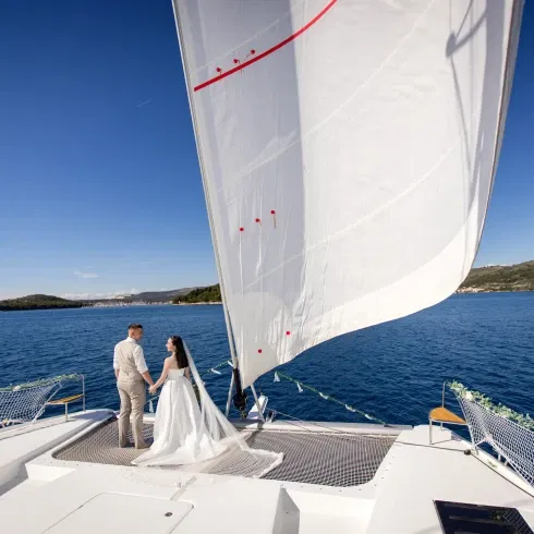 Ein Brautpaar steht Hand in Hand auf einem Segelkatamaran und blickt aufs offene Meer. Die Braut trägt ein weißes Kleid mit langem Schleier, der Bräutigam einen hellen Anzug.