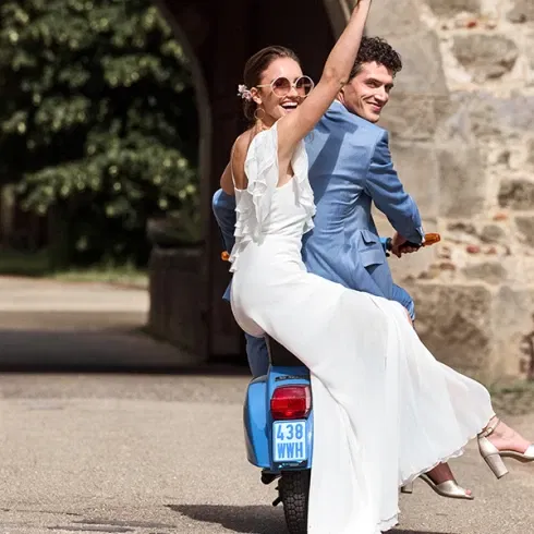 Frisch verheiratetes Paar fährt fröhlich auf einem blauen Roller davon, die Braut hält den Brautstrauß in die Luft.