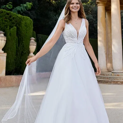 Strahlende Braut in einem romantischen A-Linien-Kleid mit tiefem V-Ausschnitt und floraler Stickerei, umgeben von mediterraner Gartenarchitektur.