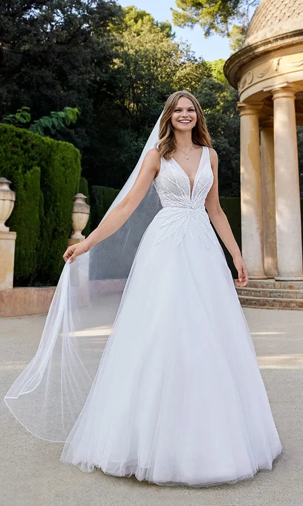 Strahlende Braut in einem romantischen A-Linien-Kleid mit tiefem V-Ausschnitt und floraler Stickerei, umgeben von mediterraner Gartenarchitektur.