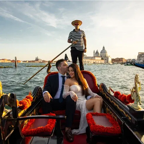 Ein verliebtes Paar in festlicher Kleidung sitzt entspannt in einer kunstvoll verzierten Gondel in Venedig. Der Gondoliere steht im Hintergrund und steuert das Boot durch das Wasser, während im Sonnenlicht historische Gebäude und die berühmte Kuppelkirche der Stadt leuchten.