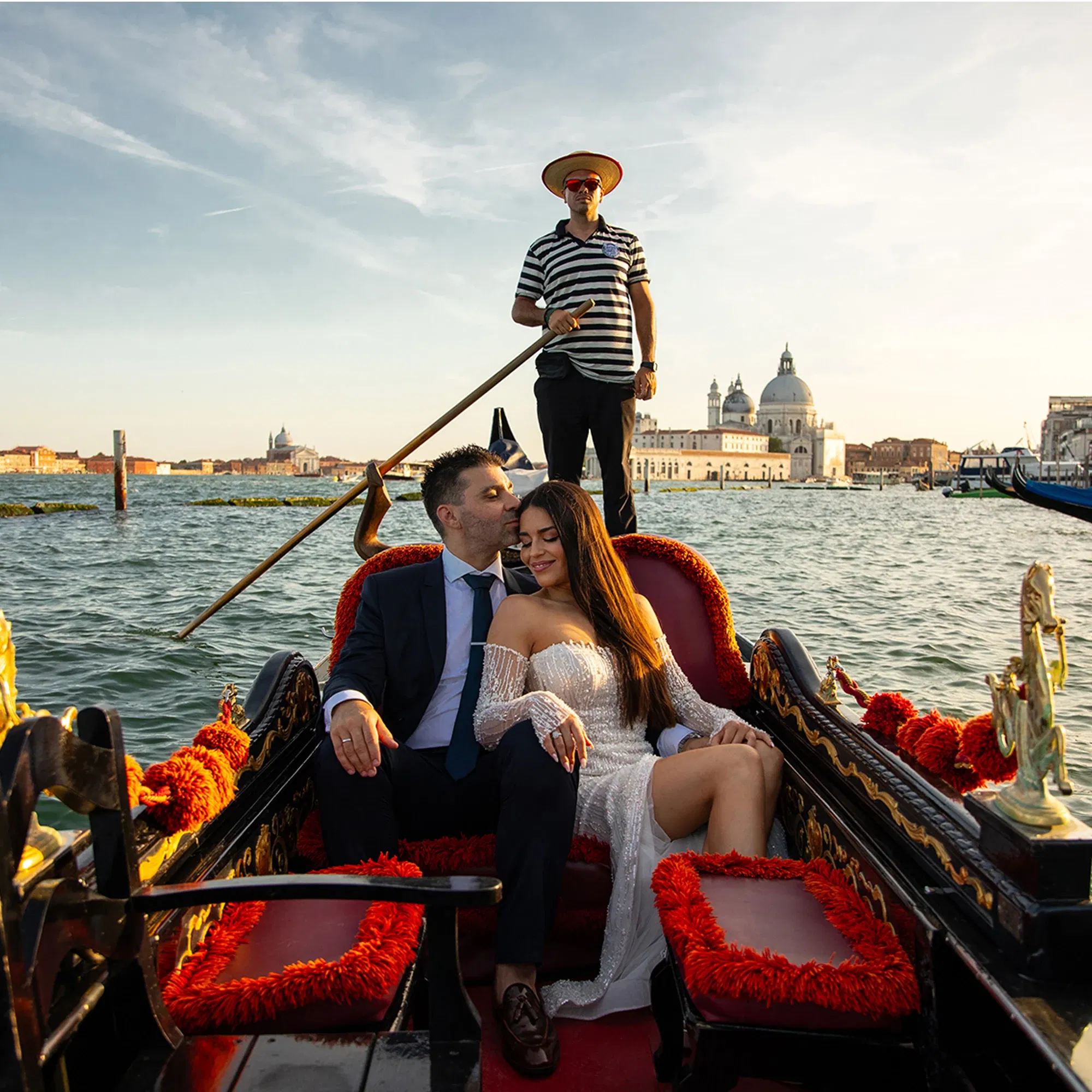 Ein verliebtes Paar in festlicher Kleidung sitzt entspannt in einer kunstvoll verzierten Gondel in Venedig. Der Gondoliere steht im Hintergrund und steuert das Boot durch das Wasser, während im Sonnenlicht historische Gebäude und die berühmte Kuppelkirche der Stadt leuchten.