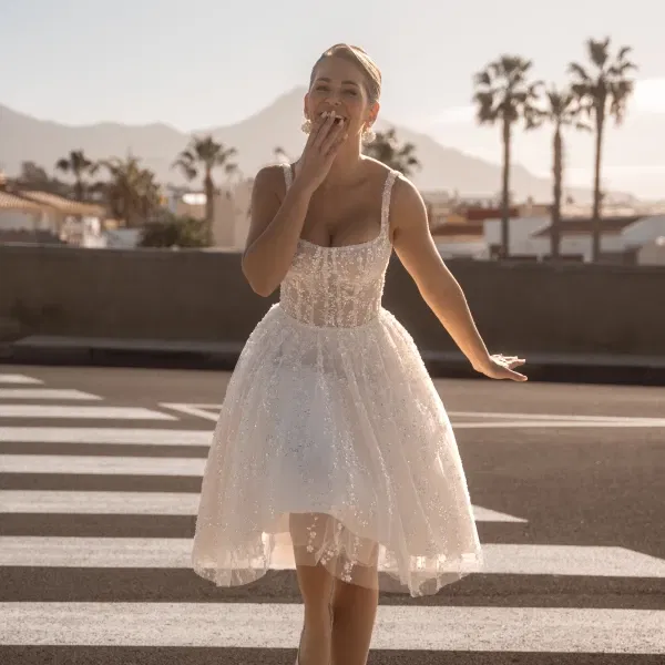 Lachende Braut in einem funkelnden, kurzen Tüllkleid mit Korsagenoberteil auf einem Zebrastreifen bei Sonnenuntergang.