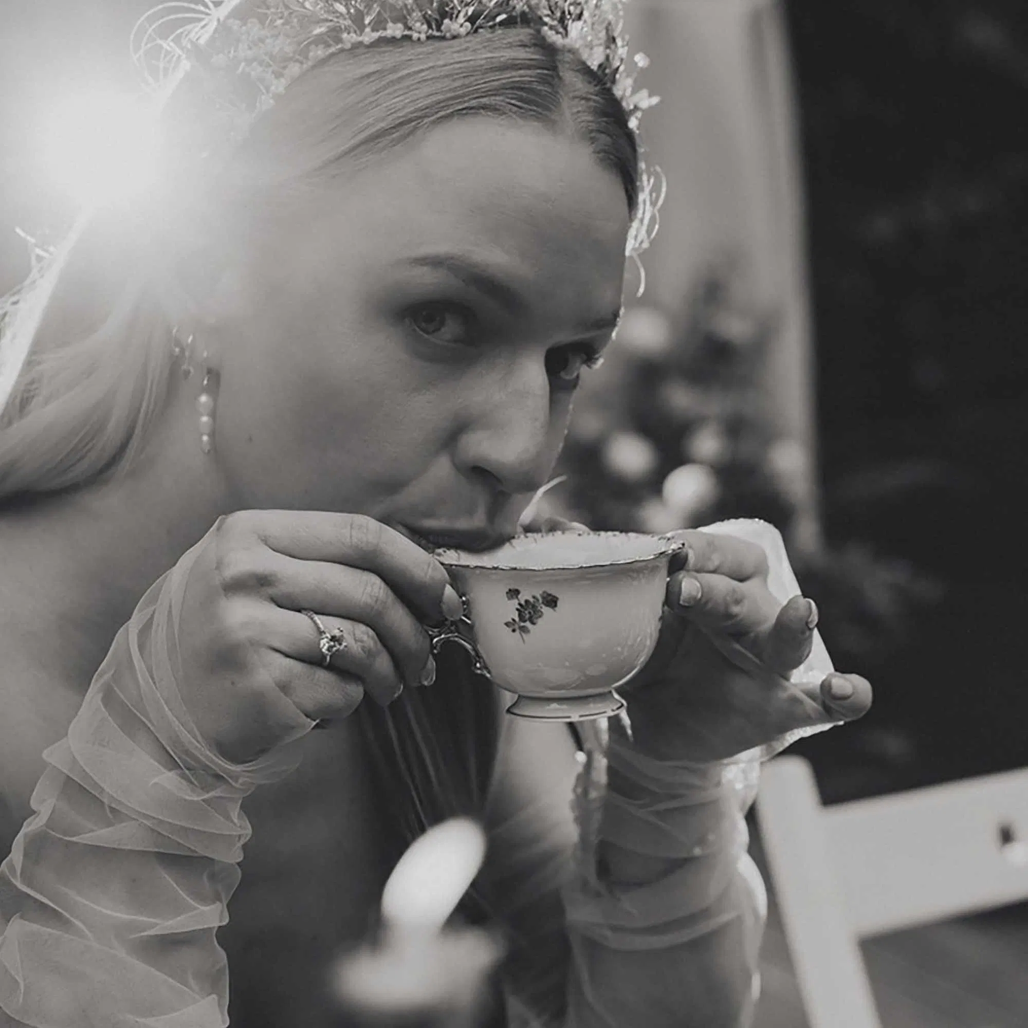 Schwarz-weißes Hochzeitsfoto einer Braut mit Diadem, die während der Feier aus einer feinen Teetasse trinkt.