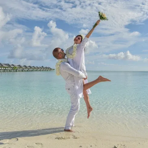 Fröhliches Hochzeitspaar feiert barfuß am Meer, der Bräutigam hebt die Braut in die Luft.