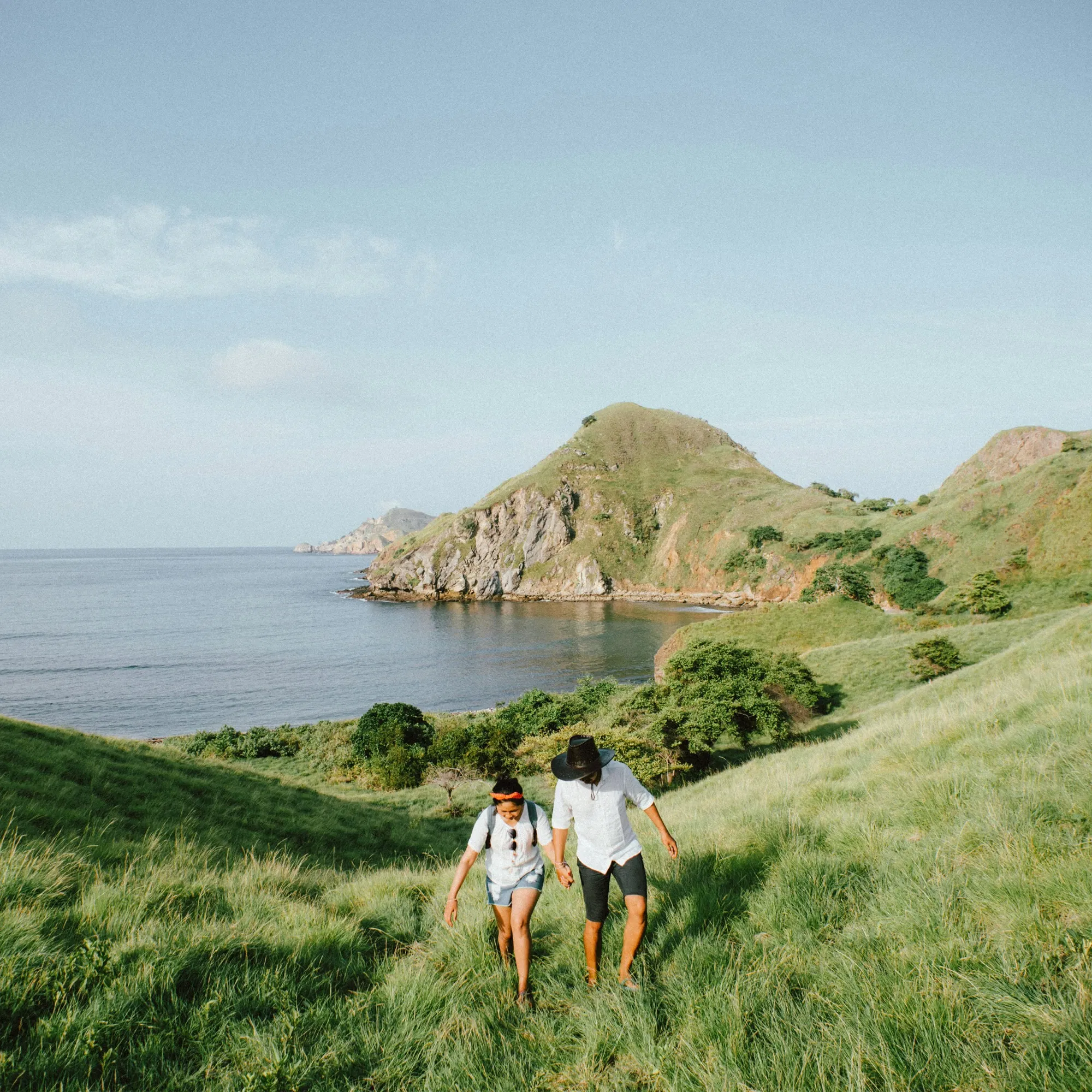 Ein Paar wandert barfuß durch sattgrünes Gras entlang einer malerischen Küste mit sanften Hügeln und ruhigem Meer im Hintergrund.