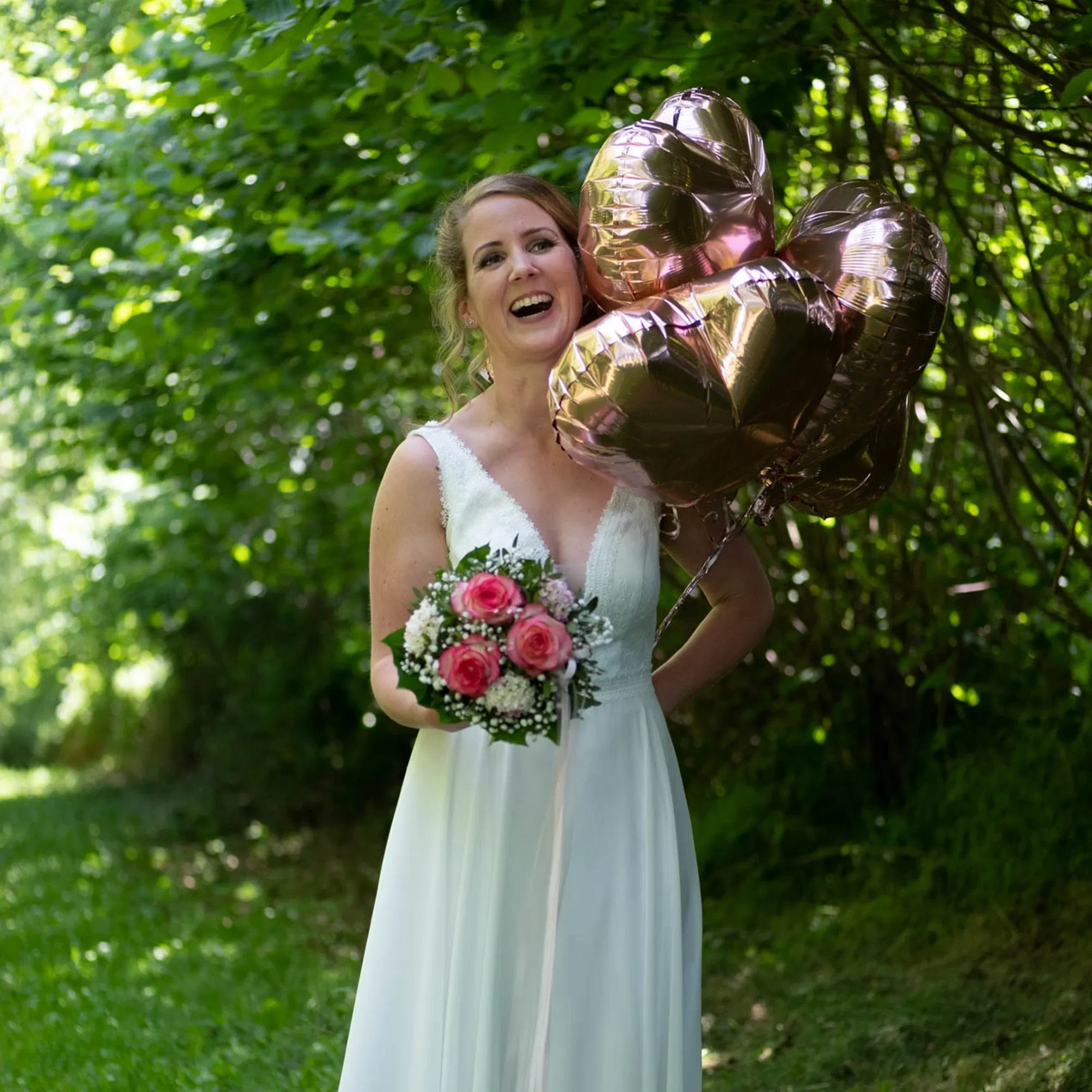 Braut in weißem Kleid lacht mit roségoldenen Herzballons und einem Strauß pinker Rosen im Grünen.