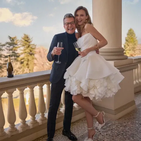 Braut in kurzem, weißen Volantkleid mit floralen Details posiert lachend mit Randy Fenoli auf einer Terrasse bei Sekt und Sonnenschein.