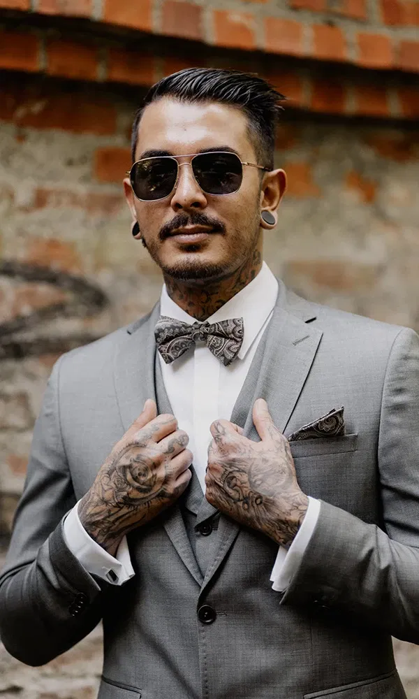 Stilvoller Bräutigam mit Tattoos, Sonnenbrille und Fliege posiert selbstbewusst.