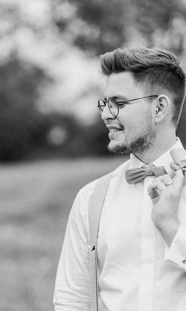 Nahaufnahme eines entspannten Bräutigams mit Brille und Fliege im Vintage-Stil.