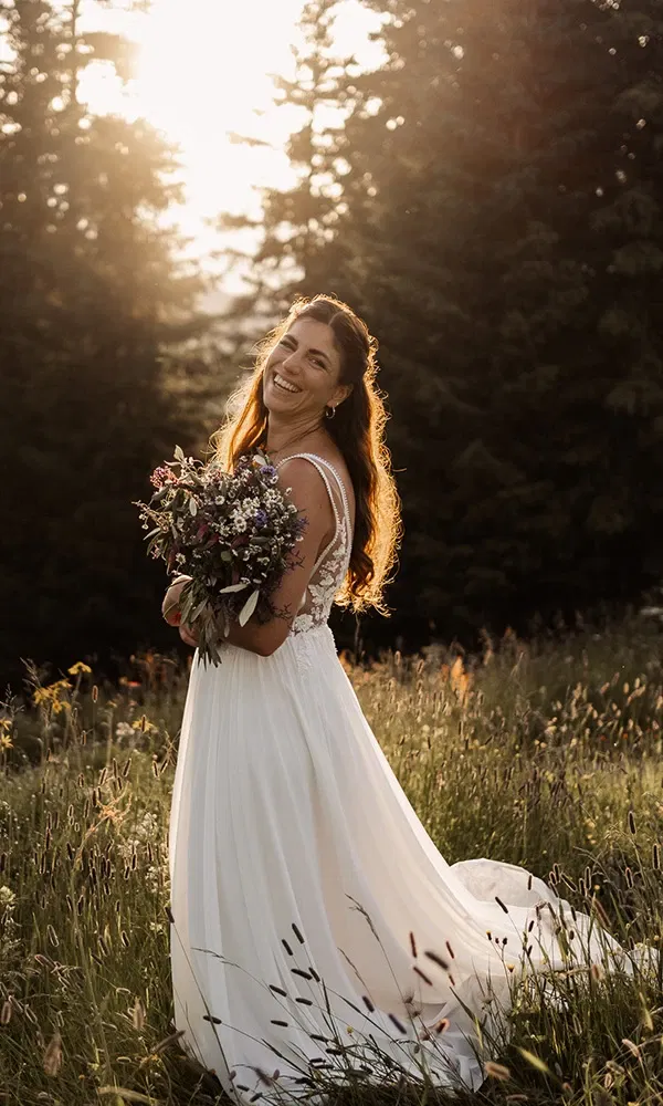 Strahlende Braut mit Blumenstrauß im Gegenlicht einer sommerlichen Wiese.