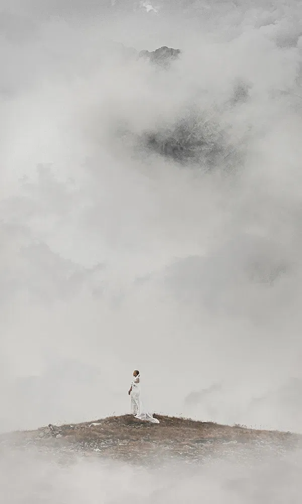 Mystische Aufnahme einer Braut im weißen Kleid auf nebliger Bergspitze.