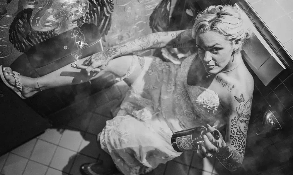 Unkonventionelle Braut raucht und trinkt in markantem Hochzeitsfoto.