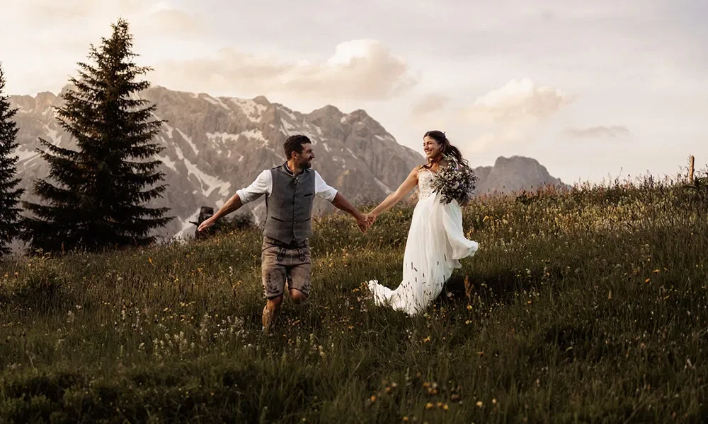 Brautpaar läuft lachend durch eine Almwiese vor beeindruckender Bergkulisse.