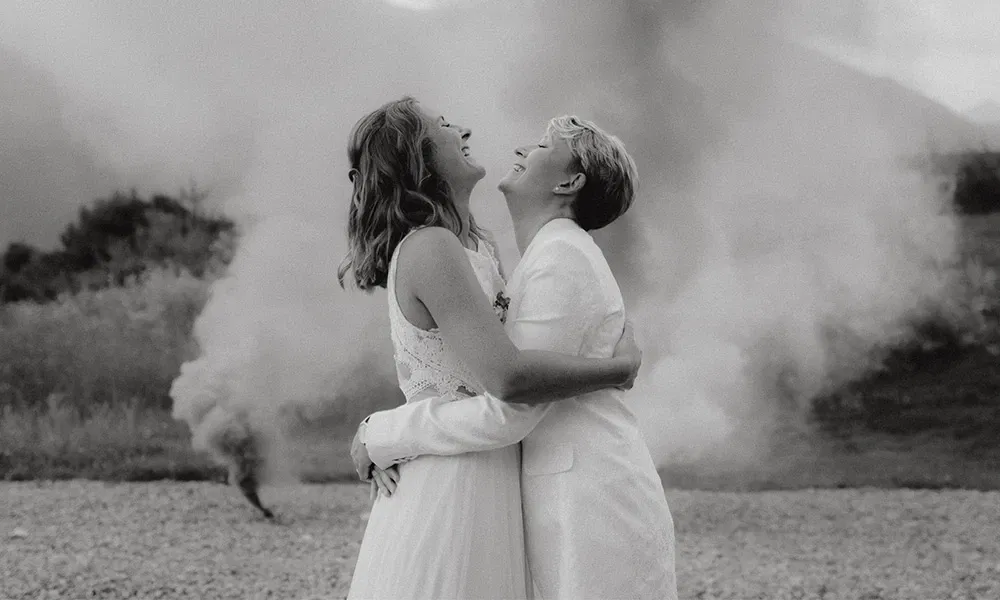 Gleichgeschlechtliches Brautpaar lacht herzlich vor aufsteigendem Rauch bei der Hochzeit.