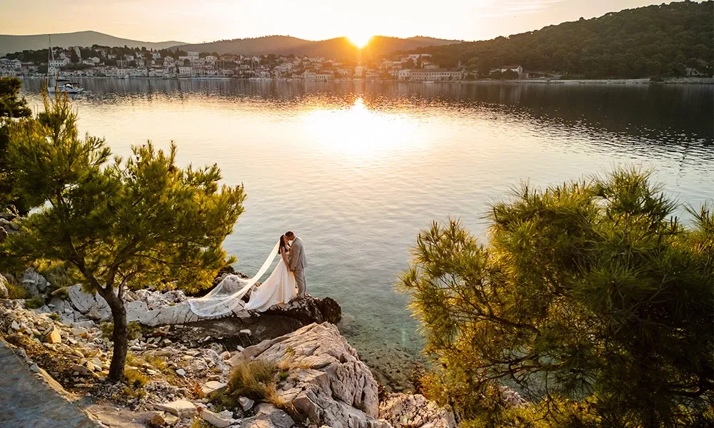 Brautpaar küsst sich auf einer Felsklippe mit Blick auf das Meer bei Sonnenuntergang.