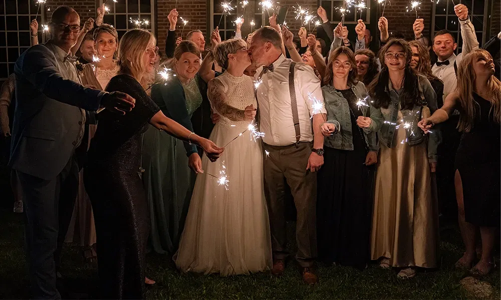 Braut und Bräutigam küssen sich umgeben von Gästen mit Wunderkerzen.