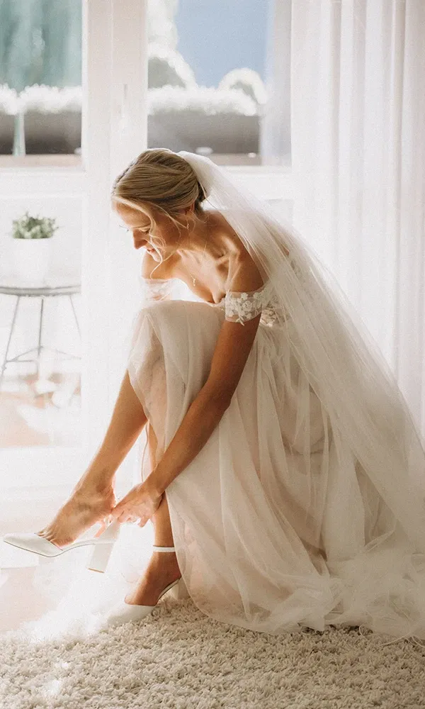 Braut sitzt im Licht und schließt den Riemen ihrer Hochzeitsschuhe.