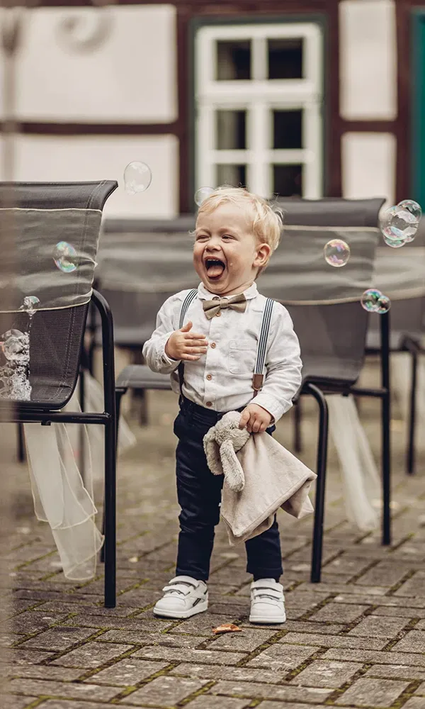 Kleiner Junge im Hochzeitsoutfit lacht ausgelassen zwischen schwebenden Seifenblasen.