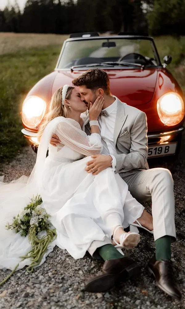 Brautpaar küsst sich vor einem Oldtimer, entspannt auf der Straße sitzend.