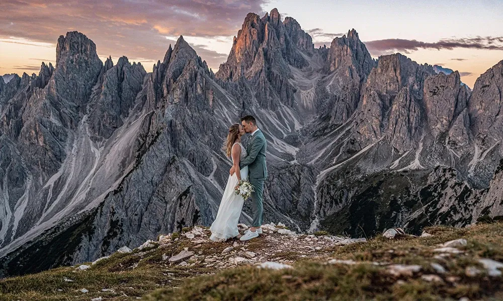 Hochzeitspaar auf Berggipfel vor dramatischer Dolomiten-Kulisse im Abendlicht.